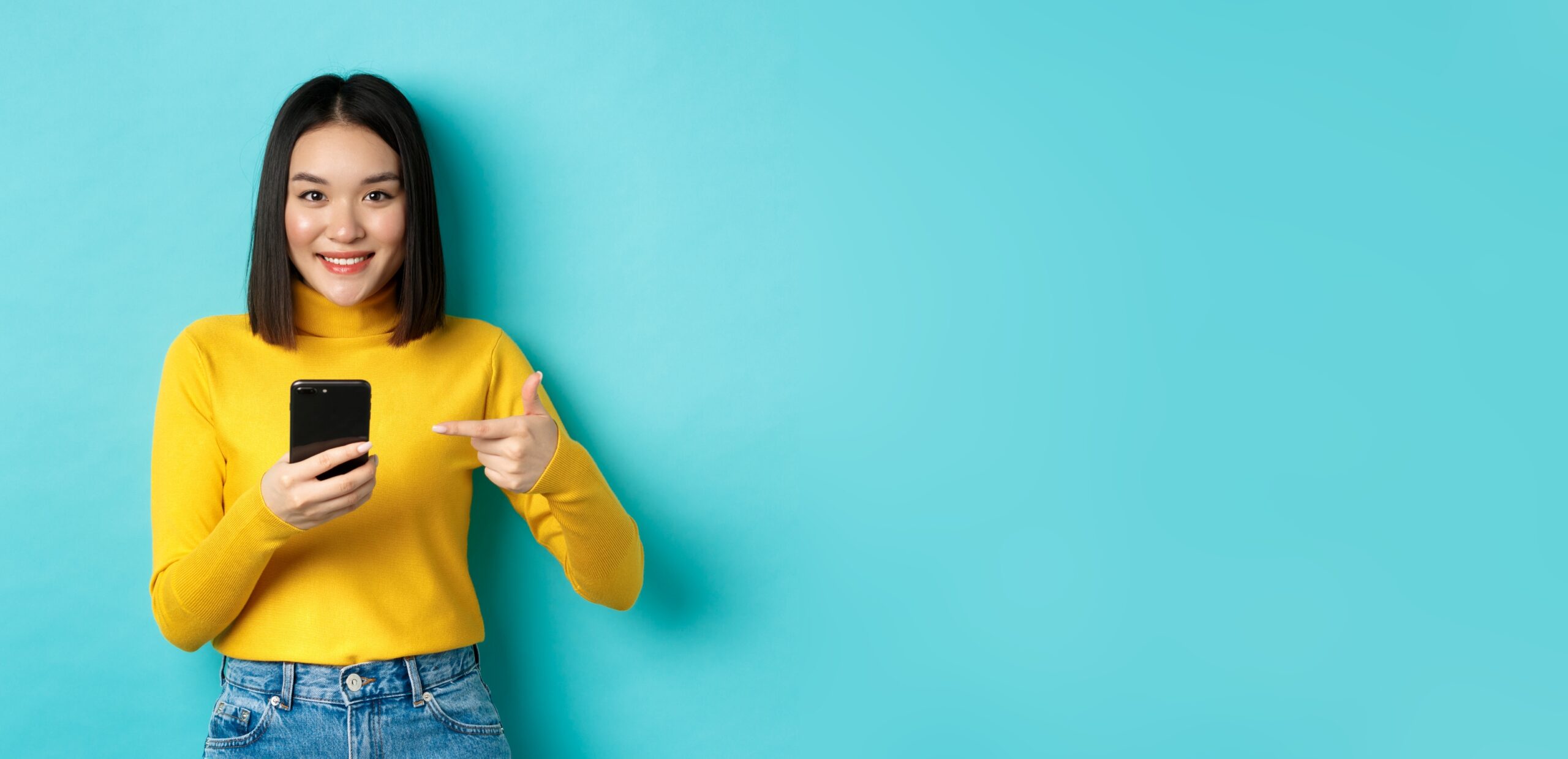 E-commerce and online shopping concept. Cute asian woman in yellow sweater pointing at smartphone, smiling at camera, standing over blue background.