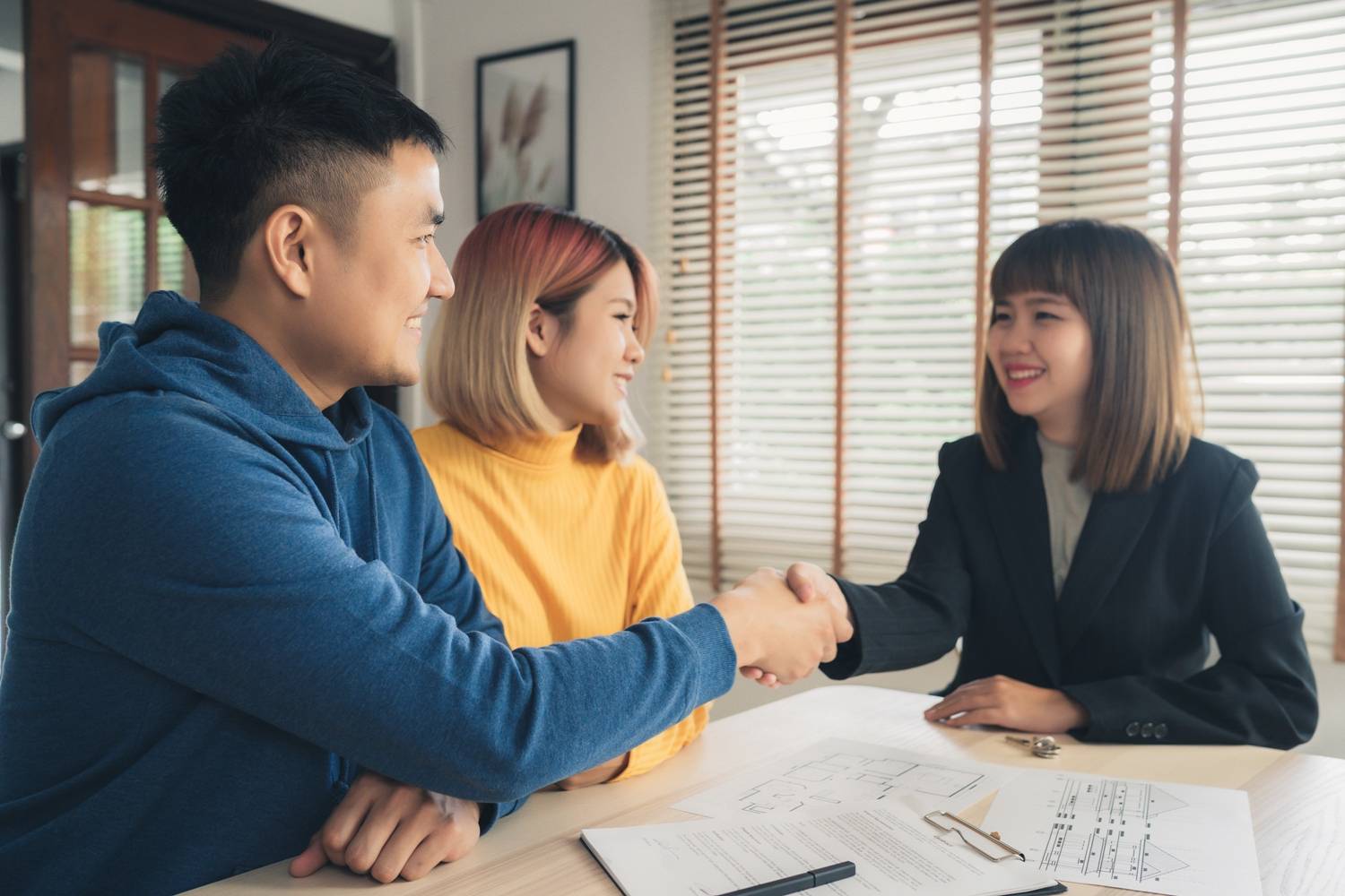 Happy young Asian couple and realtor agent. Cheerful young man signing some documents and handshaking with broker while sitting at desk. Signing good condition contract.