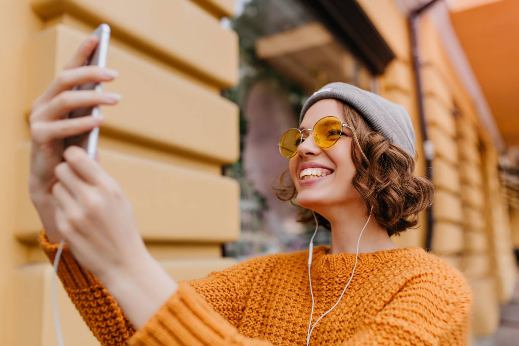 Lovely white girl with curly hairstyle making selfie in new outfit enjoying good weather. Outdoor portrait of ecstatic young woman in trendy sweater taking picture of herself beside old building.