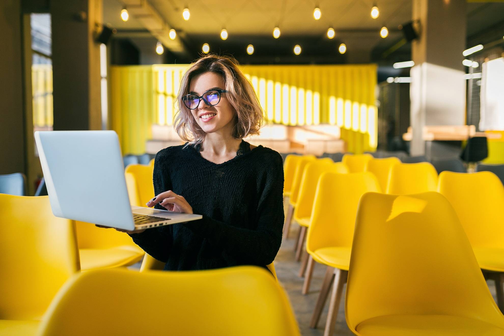 portrait of young attractive woman sitting in lecture hall working on laptop wearing glasses, student learning in classroom with many yellow chairs